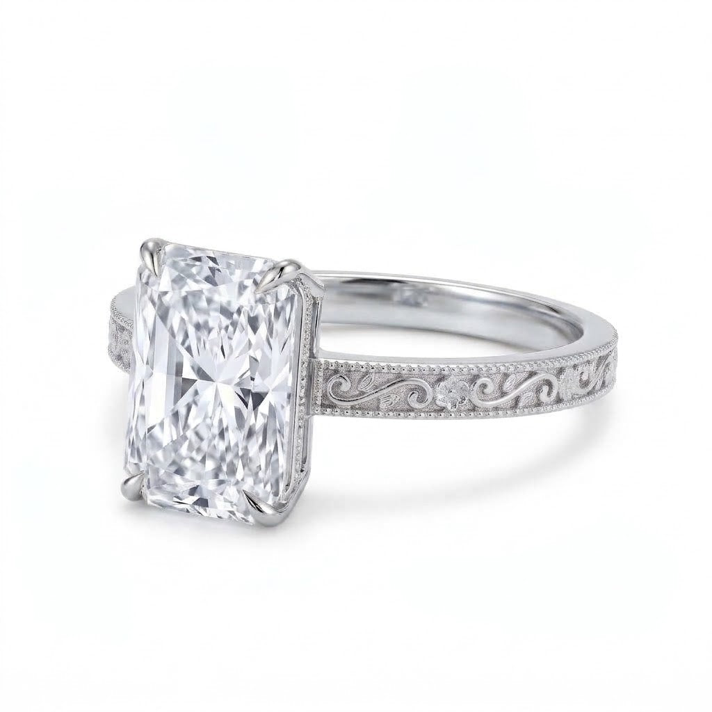 Radiant-cut solitaire engagement ring with four claw prongs, floral scroll engravings and milgrain borders on a white-metal band