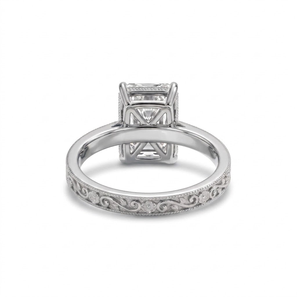 Radiant-cut solitaire engagement ring with four claw prongs, floral scroll engravings and milgrain borders on a white-metal band